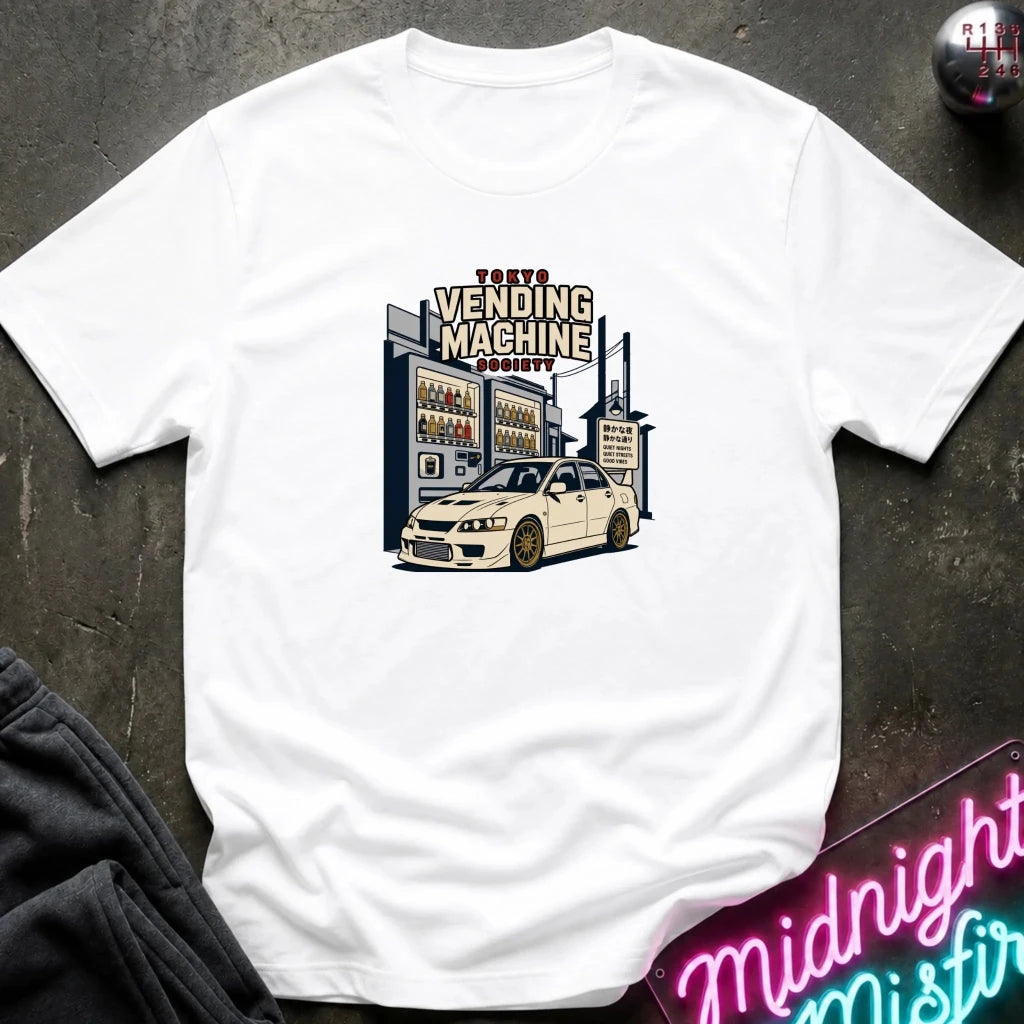 A white short-sleeve graphic t-shirt laid flat on a dark textured surface. The graphic features a vintage-style illustration of a 'Tokyo Vending Machine Society' building with a white sports car parked in front.