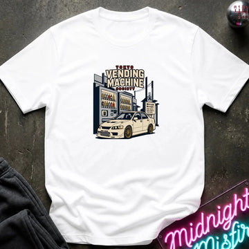A white short-sleeve graphic t-shirt laid flat on a dark textured surface. The graphic features a vintage-style illustration of a 'Tokyo Vending Machine Society' building with a white sports car parked in front.