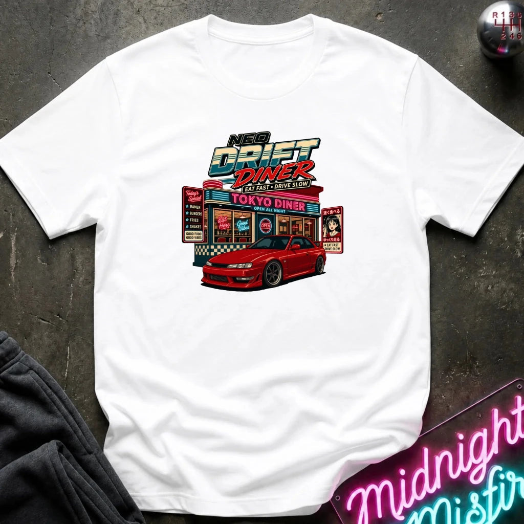 A white short-sleeve t-shirt laid flat on a dark textured surface, featuring a colorful graphic print of a red sports car parked in front of a retro 'Neon Drift Diner' sign. A neon sign and dark clothing are partially visible as styled props.
