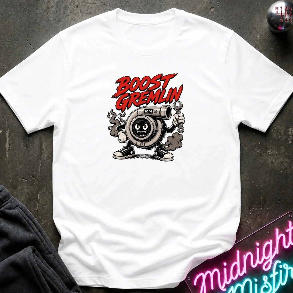 A white short-sleeve graphic T-shirt laid flat on a dark textured surface. The shirt features a cartoon turbocharger character with the words 'BOOST GREMLIN' in red above it.