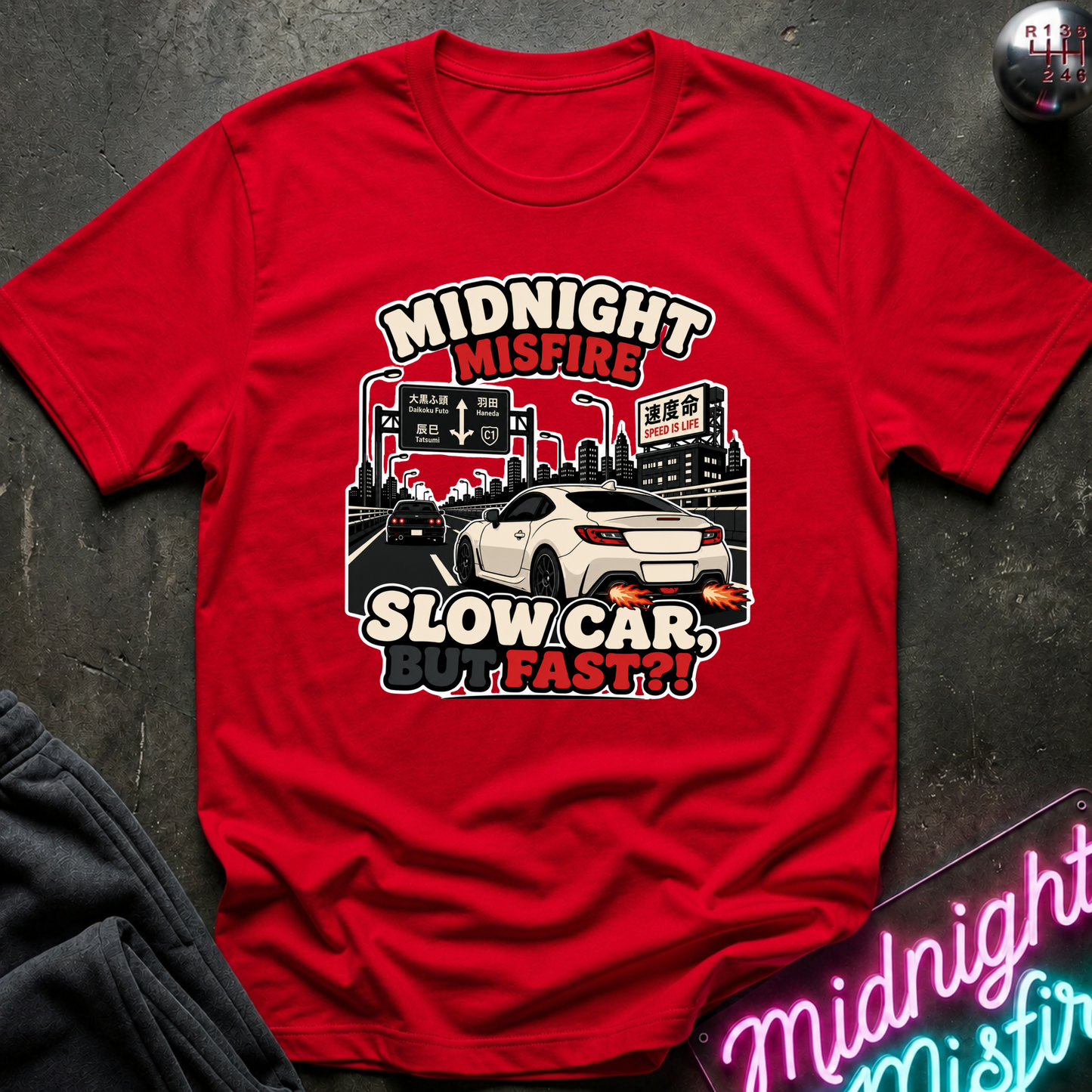 A red short-sleeve graphic T-shirt laid flat on a dark textured surface. The shirt features a large front print with the words “MIDNIGHT MISFIRE” and “SLOW CAR, BUT FAST??” in white and red lettering, with a white sports car and city street scene illustration.