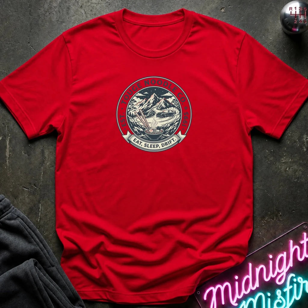A red short-sleeve crewneck t-shirt laid flat on a dark textured surface. The center of the shirt features a circular graphic with a mountain illustration and the words 