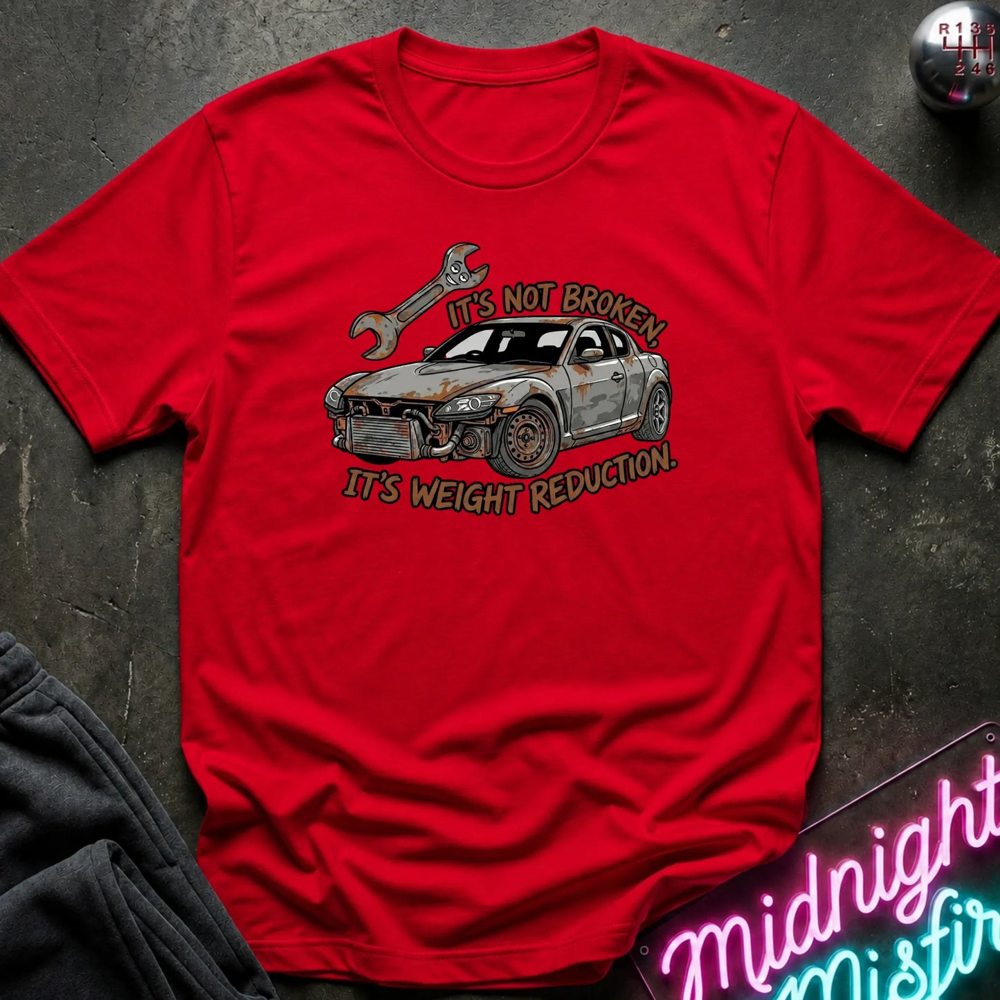 A red short-sleeve graphic T-shirt laid flat on a dark textured surface. The shirt features a distressed illustration of a damaged gray sports car with a wrench above it and the text 'IT'S NOT BROKEN.