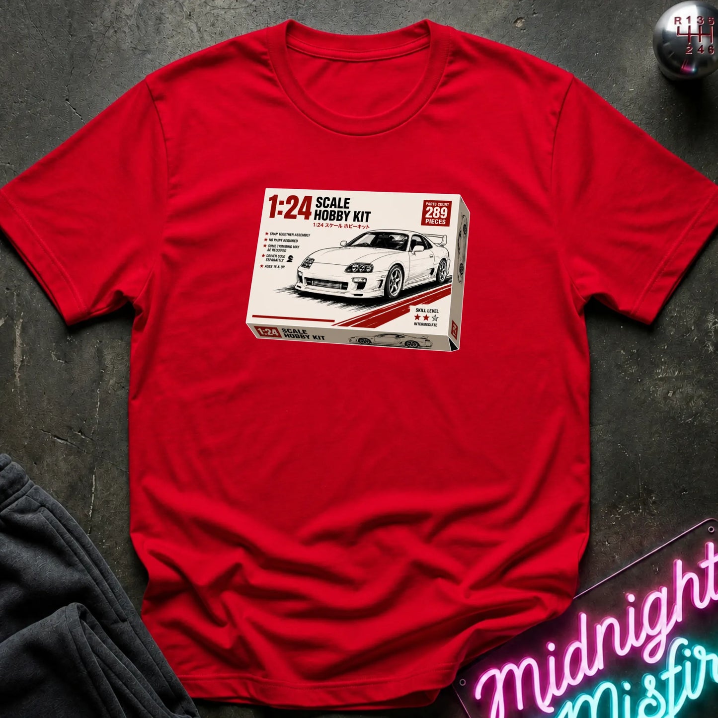 A red short-sleeve t-shirt laid flat on a dark textured surface, featuring a printed graphic of a 1:24 scale model kit box for a white sports car. A neon sign is partially visible in the bottom right corner.