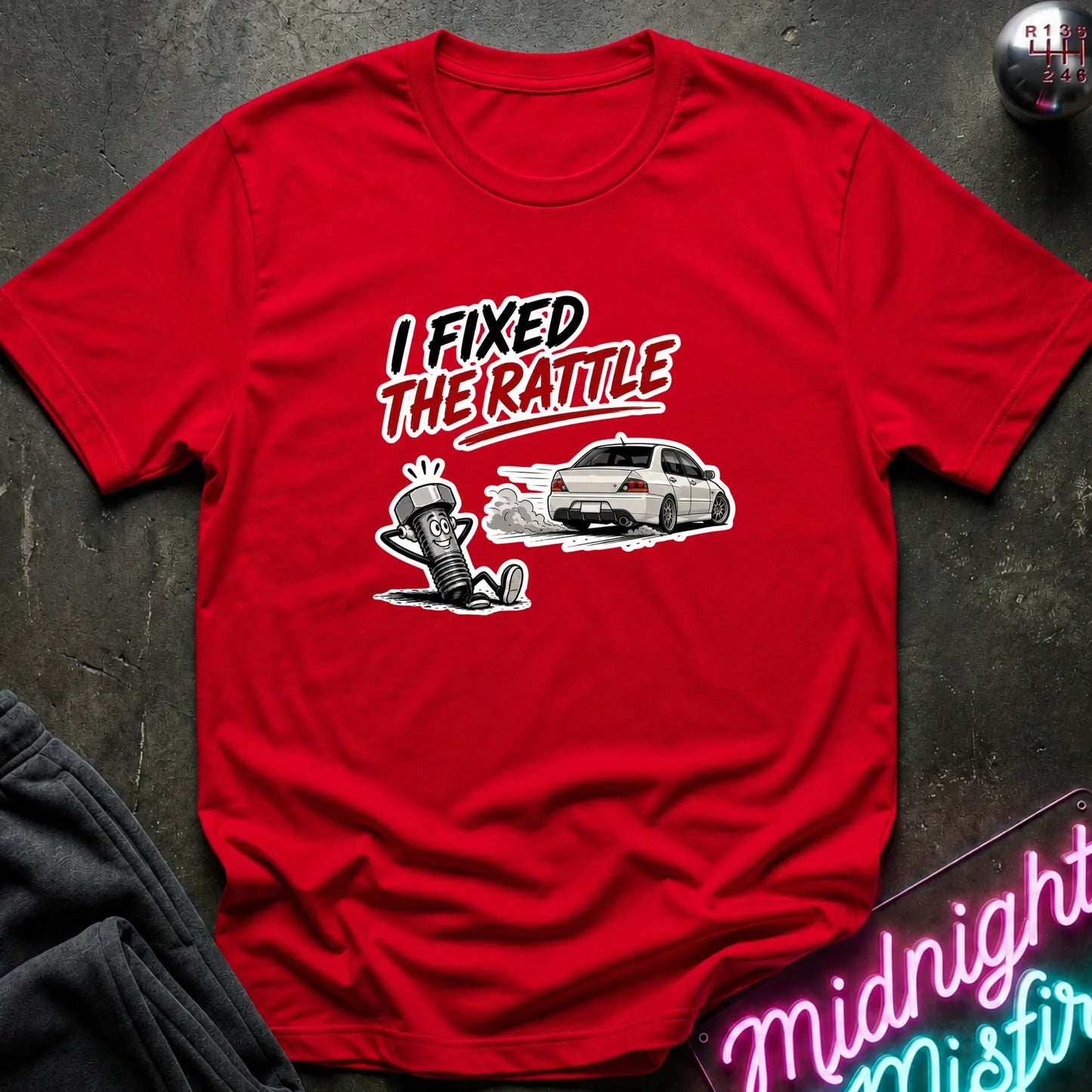 A red short-sleeve graphic t-shirt laid flat on a dark textured surface. The graphic features the text 'I FIXED THE RATTLE' in white and black, above an illustration of a white car and a cartoon character.