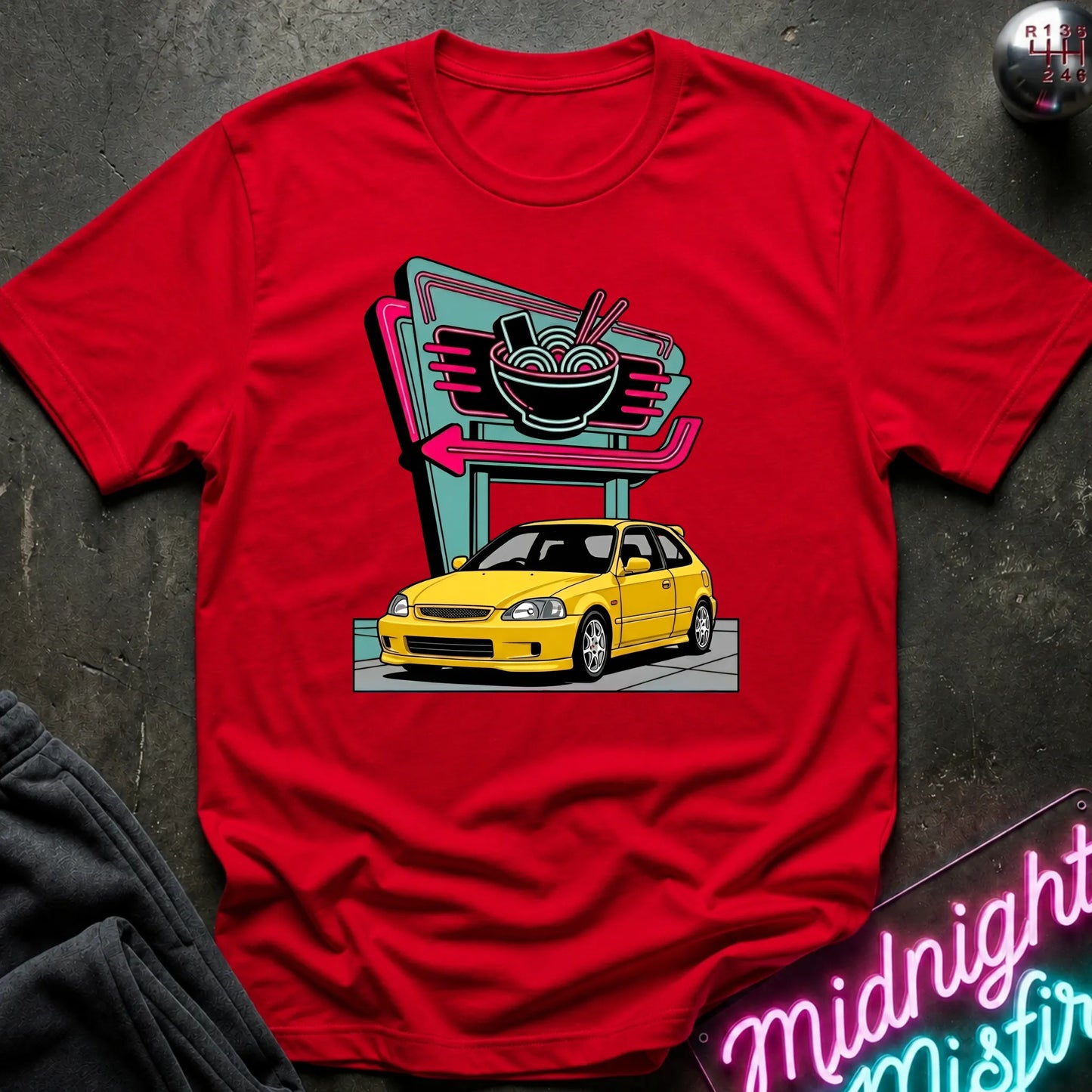 A red short-sleeve graphic t-shirt laid flat on a dark textured surface. The graphic shows a yellow hatchback car parked in front of a retro neon ramen shop sign.