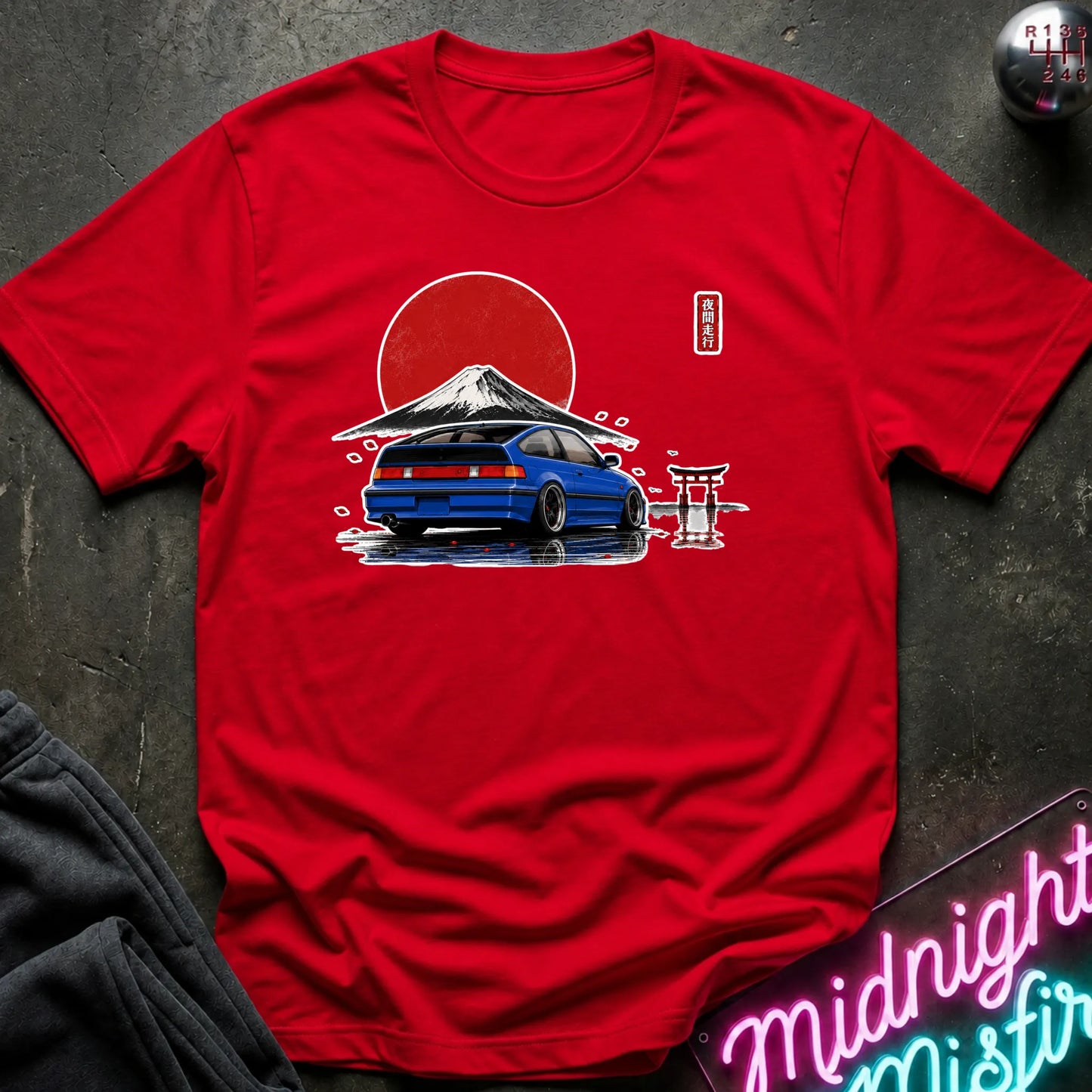 A red short-sleeve graphic T-shirt laid flat on a dark textured surface. The shirt features a printed design of a blue car in front of a red sun and mountain, with a small Japanese torii gate and vertical text on the chest.
