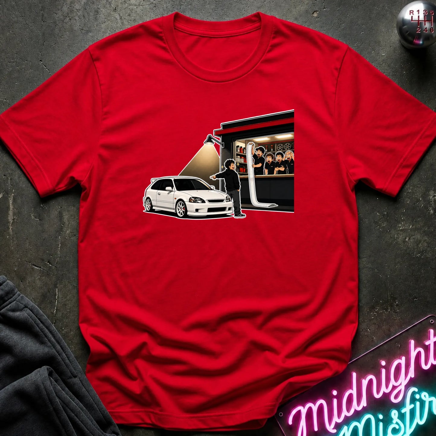 A red short-sleeve crewneck t-shirt featuring a graphic print of a white hatchback car and a person in front of a building, laid flat on a dark textured surface with a neon sign and dark sweatpants partially visible.