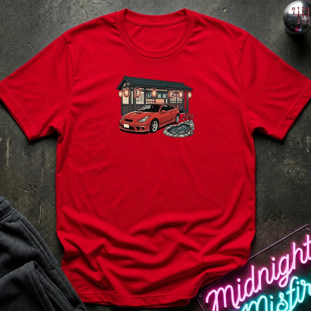 A red short-sleeve crewneck t-shirt laid flat on a dark textured surface, featuring a graphic print of a red sports car parked in front of a Japanese-style building. 
