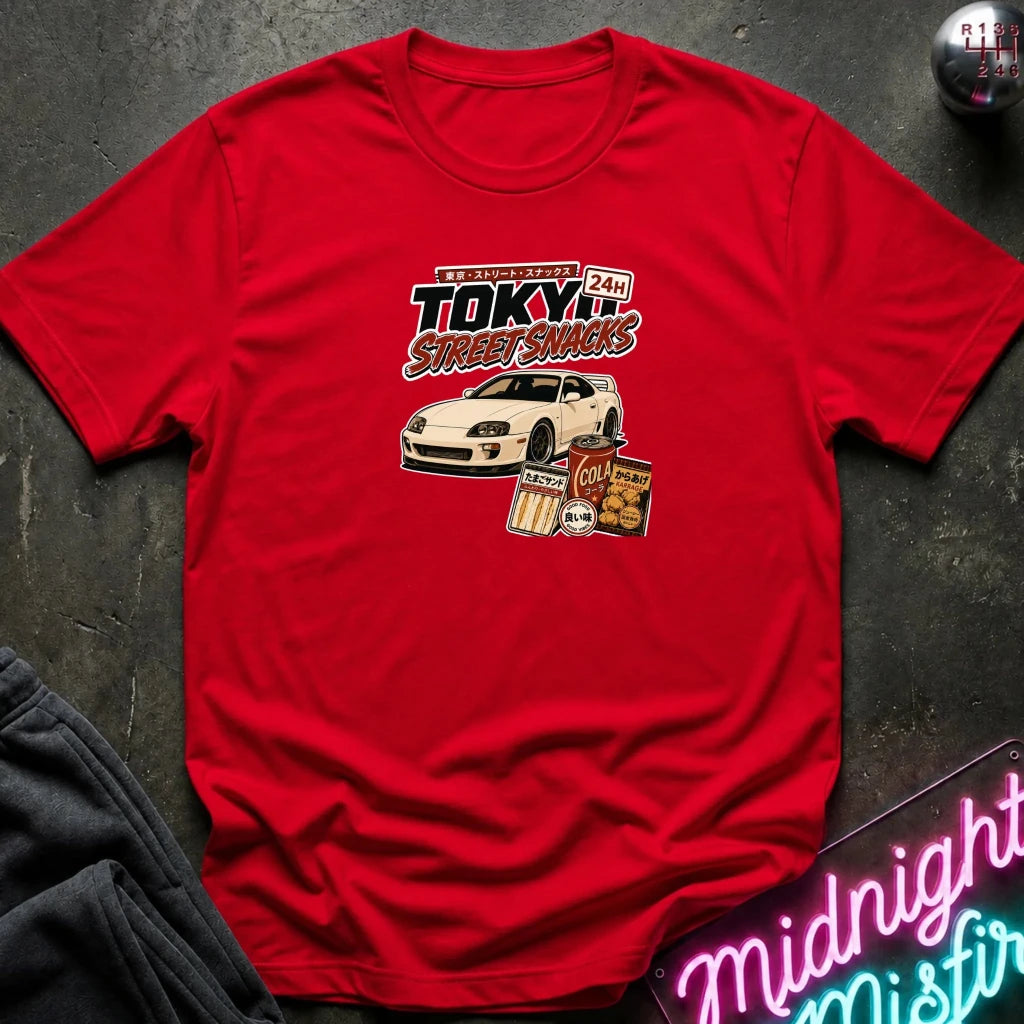 A red short-sleeve graphic T-shirt laid flat on a dark textured surface. The shirt features a central graphic with the text 'Tokyo Street Snacks' above a white sports car and snack illustrations.