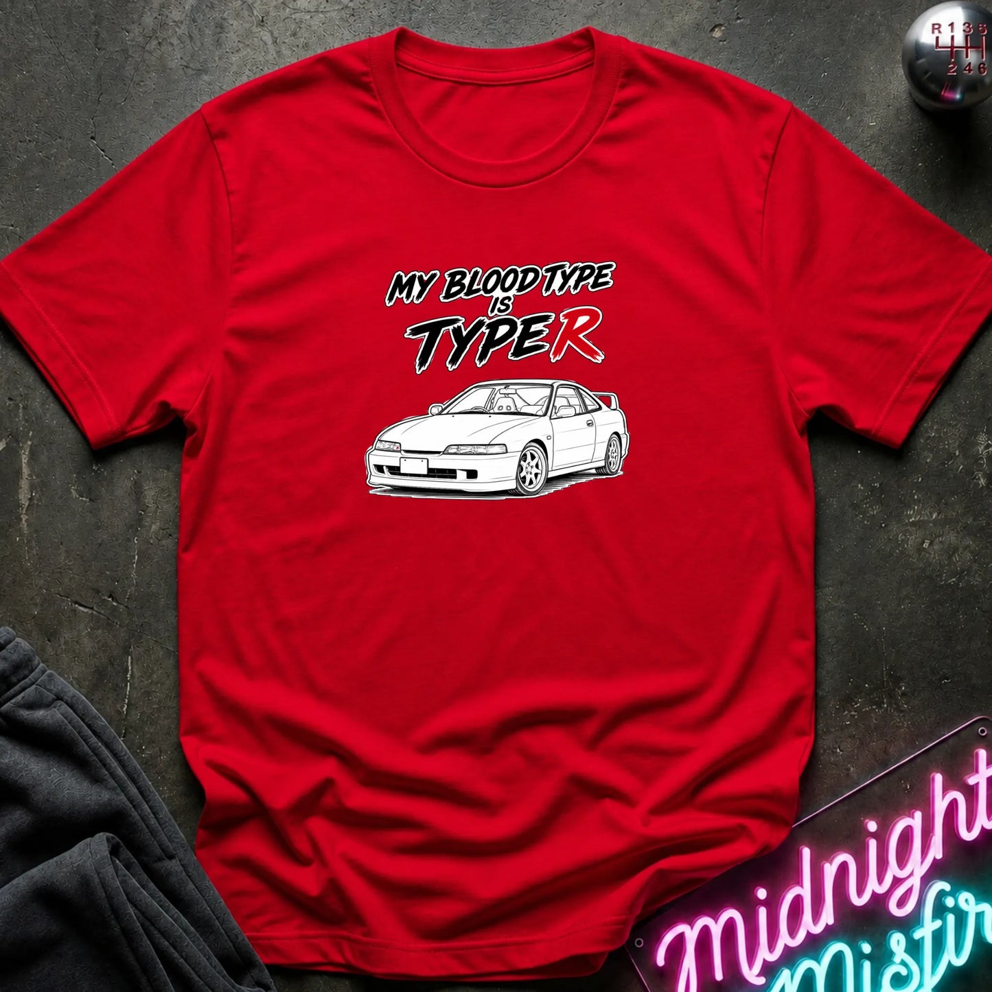 A red short-sleeve graphic T-shirt laid flat on a dark textured surface. The shirt features a white car illustration and the text 'MY BLOOD TYPE IS TYPE R' in black and red.