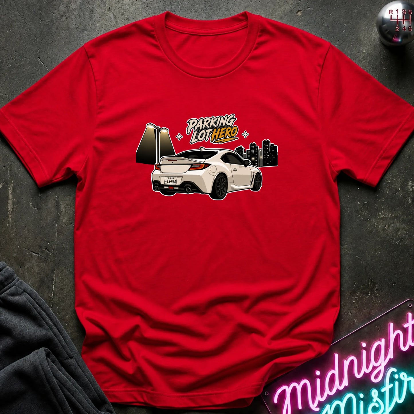 A red short-sleeve crewneck t-shirt laid flat on a dark textured surface. The shirt features a graphic print of a white sports car with the text 'PARKING LOT HERO' in yellow and white, alongside a city skyline.