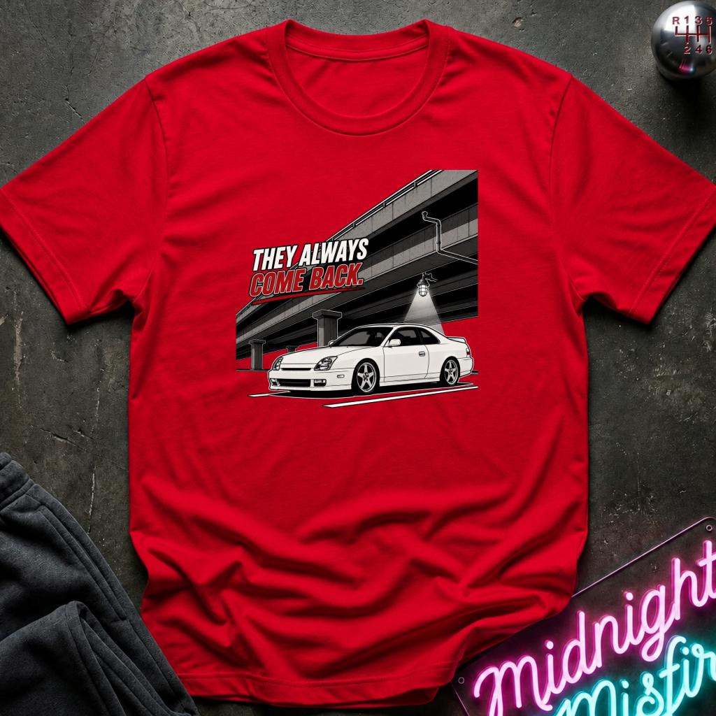 A red short-sleeve crewneck t-shirt laid flat on a dark textured surface. The shirt features a graphic print of a white sports car under an overpass with the text 'THEY ALWAYS COME BACK' in white and red.