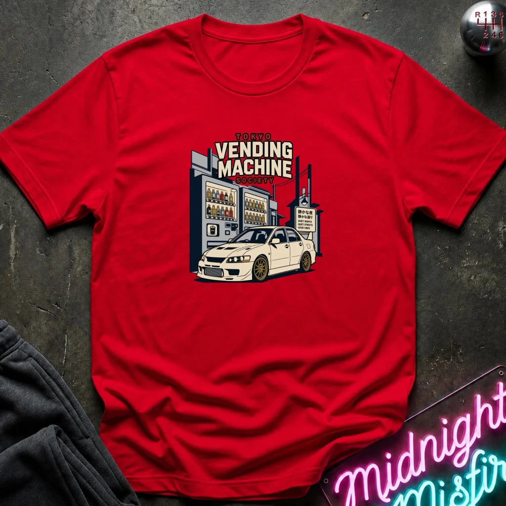 A red short-sleeve crewneck t-shirt laid flat on a dark textured surface, featuring a graphic print of a white sports car in front of a vending machine with the text 'VENDING MACHINE'.