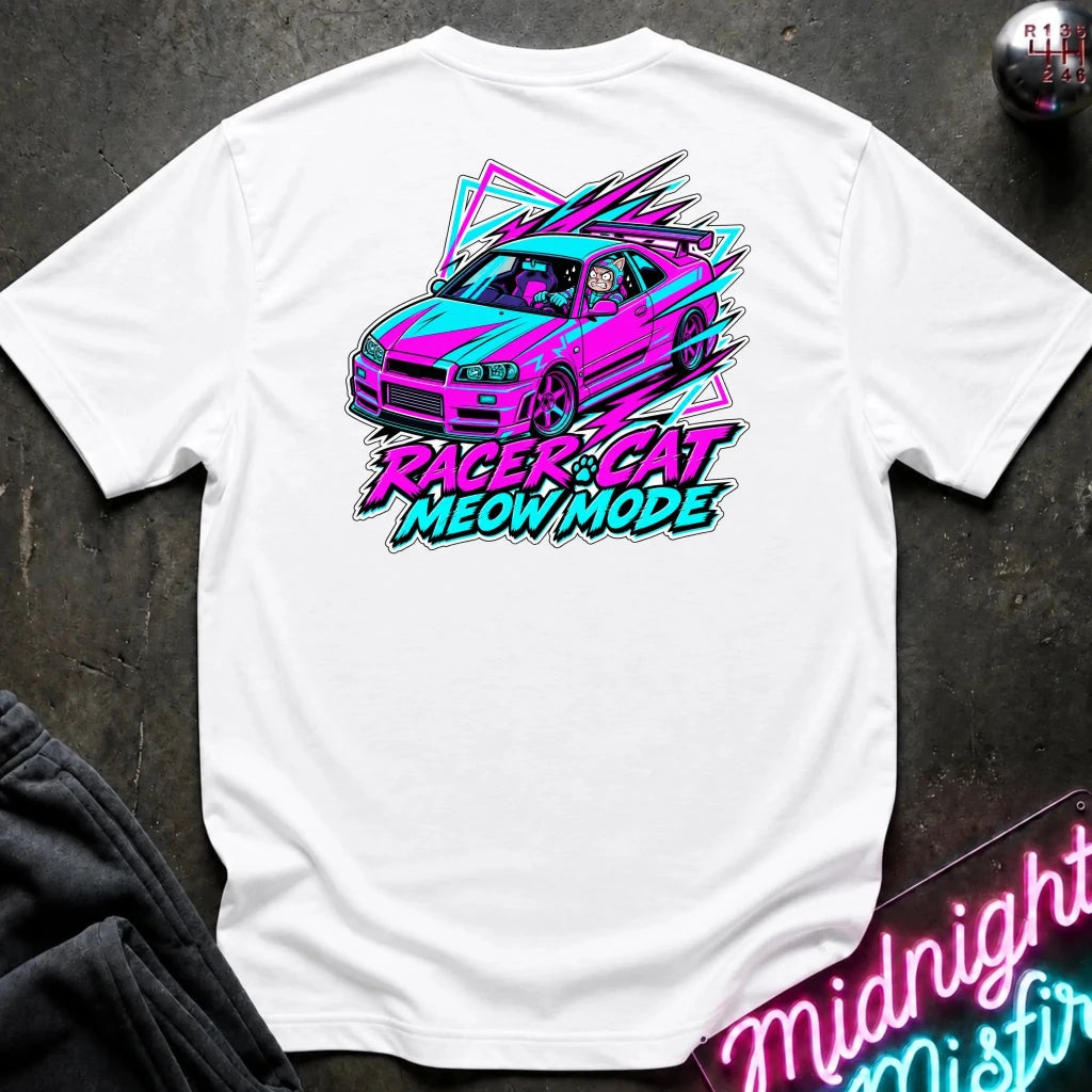A white short-sleeve t-shirt laid flat on a dark textured surface, featuring a colorful graphic of a pink and blue race car with the text 'RACER CAT MEOW MODE' in neon-style lettering. A gear shift knob and a neon sign are partially visible as props.