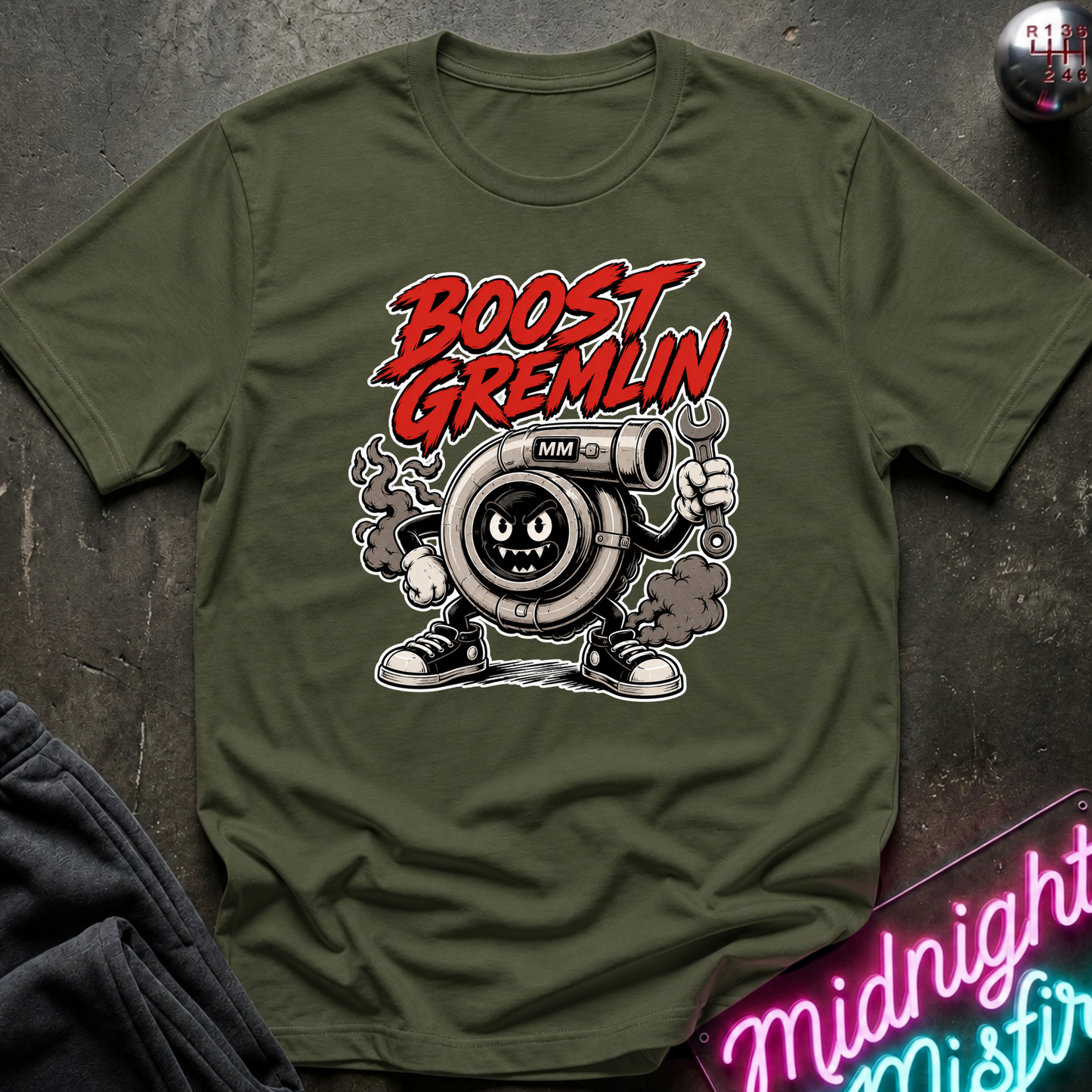 An olive green graphic t-shirt featuring a cartoon turbocharger character with the text 'BOOST GREMLIN' in red and white. The shirt is laid flat on a dark textured surface with a neon sign and dark pants partially visible in the corners.