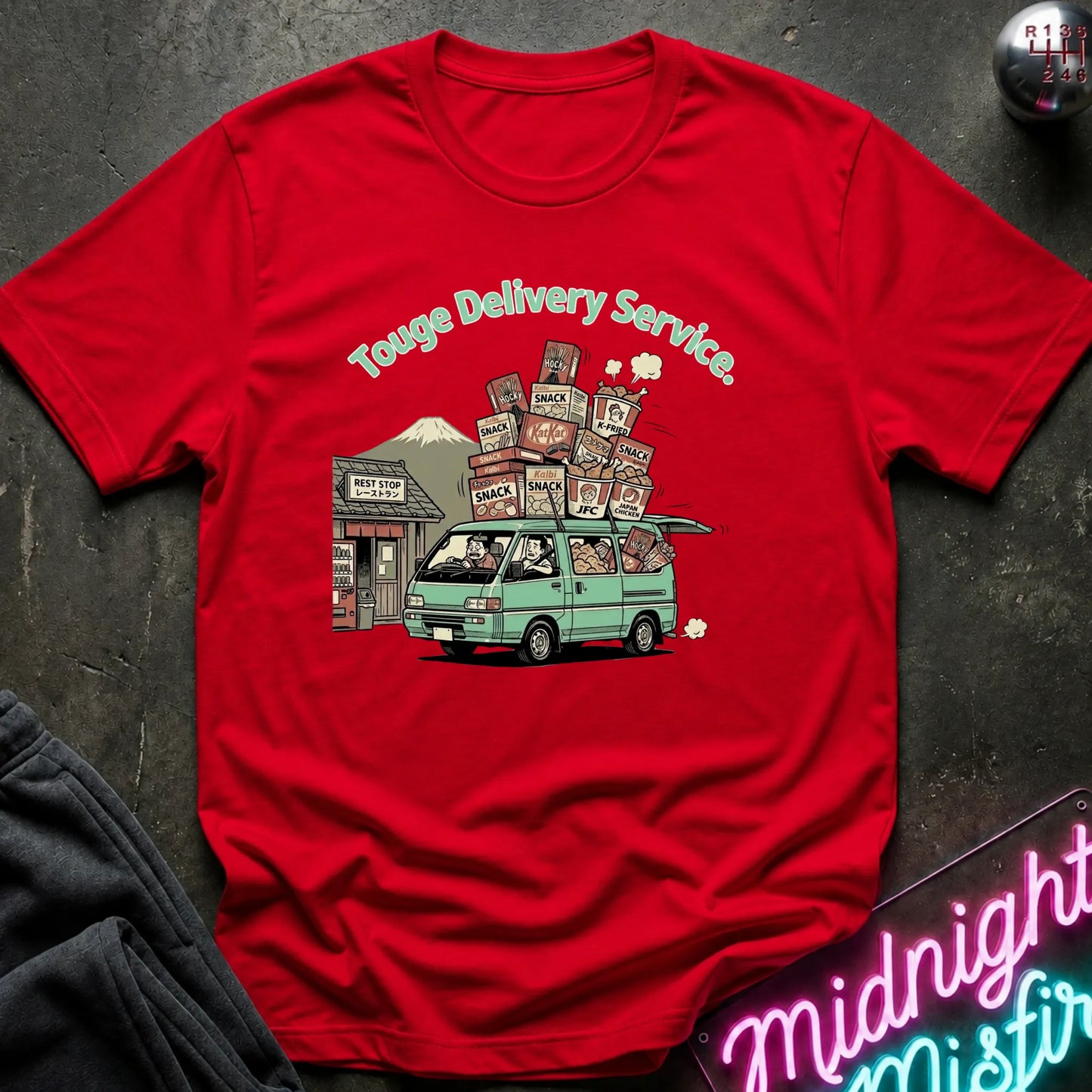 A red short-sleeve graphic t-shirt laid flat on a dark textured surface. The graphic shows a light green van carrying stacked packages with the text 'Touge Delivery Service.