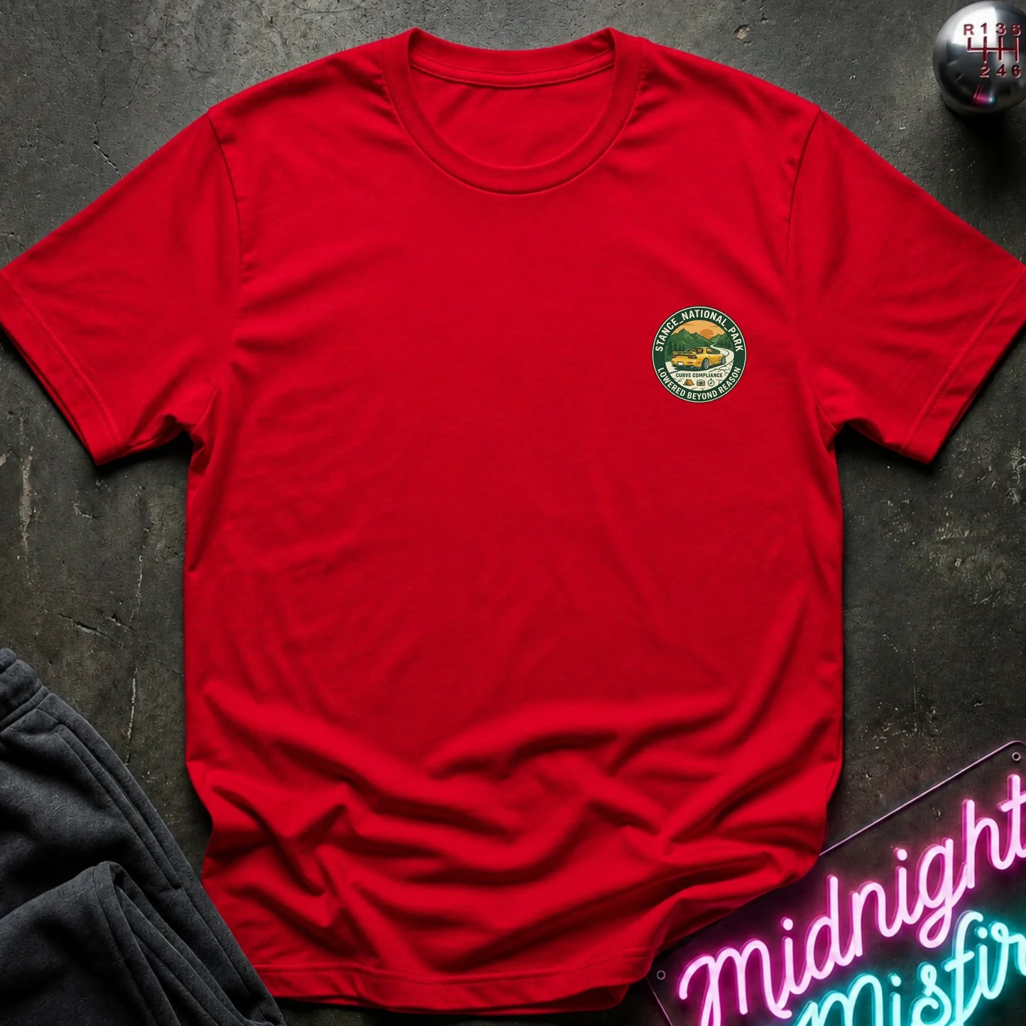 A bright crewneck t-shirt with a circular logo on the left chest, laid flat on a dark textured surface with a neon sign and dark pants in the background.