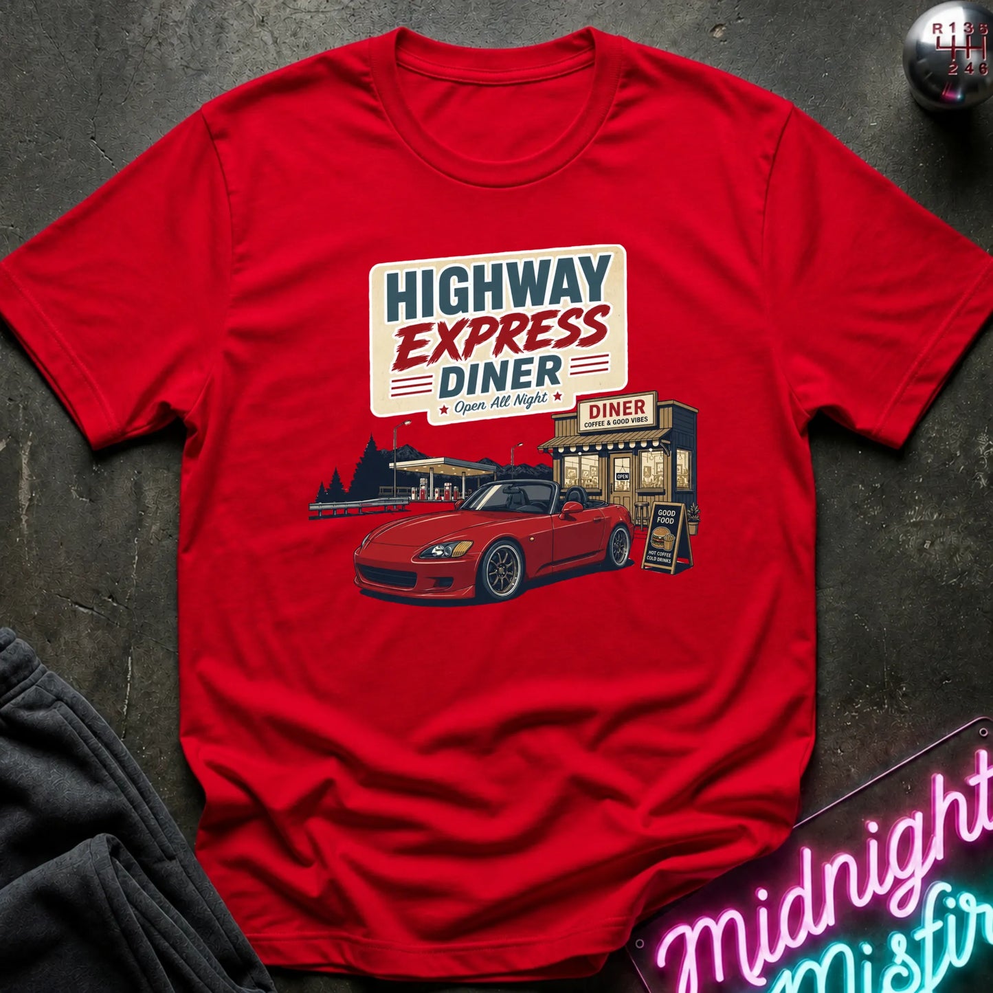 A red short-sleeve graphic T-shirt laid flat on a dark textured surface. The shirt features a retro-style diner illustration with the text 'HIGHWAY EXPRESS DINER' and a red sports car.