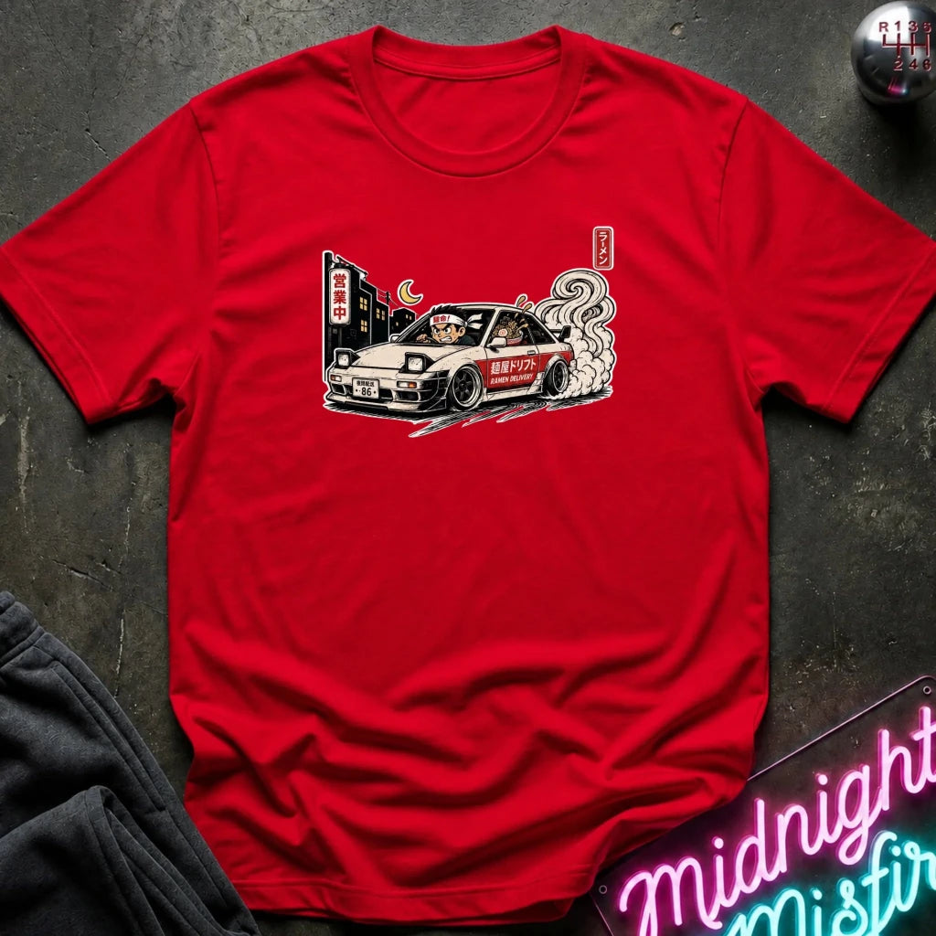A red short-sleeve crewneck t-shirt laid flat on a dark textured surface, featuring a graphic print of a white sports car with smoke and Japanese-style signs. A folded pair of dark pants is visible in the lower-left corner, and a neon sign and a small round object appear in the lower-right and upper-right corners.