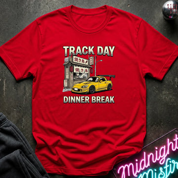 Red short-sleeve graphic T-shirt laid flat on a dark textured surface. The shirt features a printed design with the words 'TRACK DAY' and 'DINNER BREAK' in white and yellow, surrounding an illustration of a yellow sports car parked outside a Japanese-style restaurant.