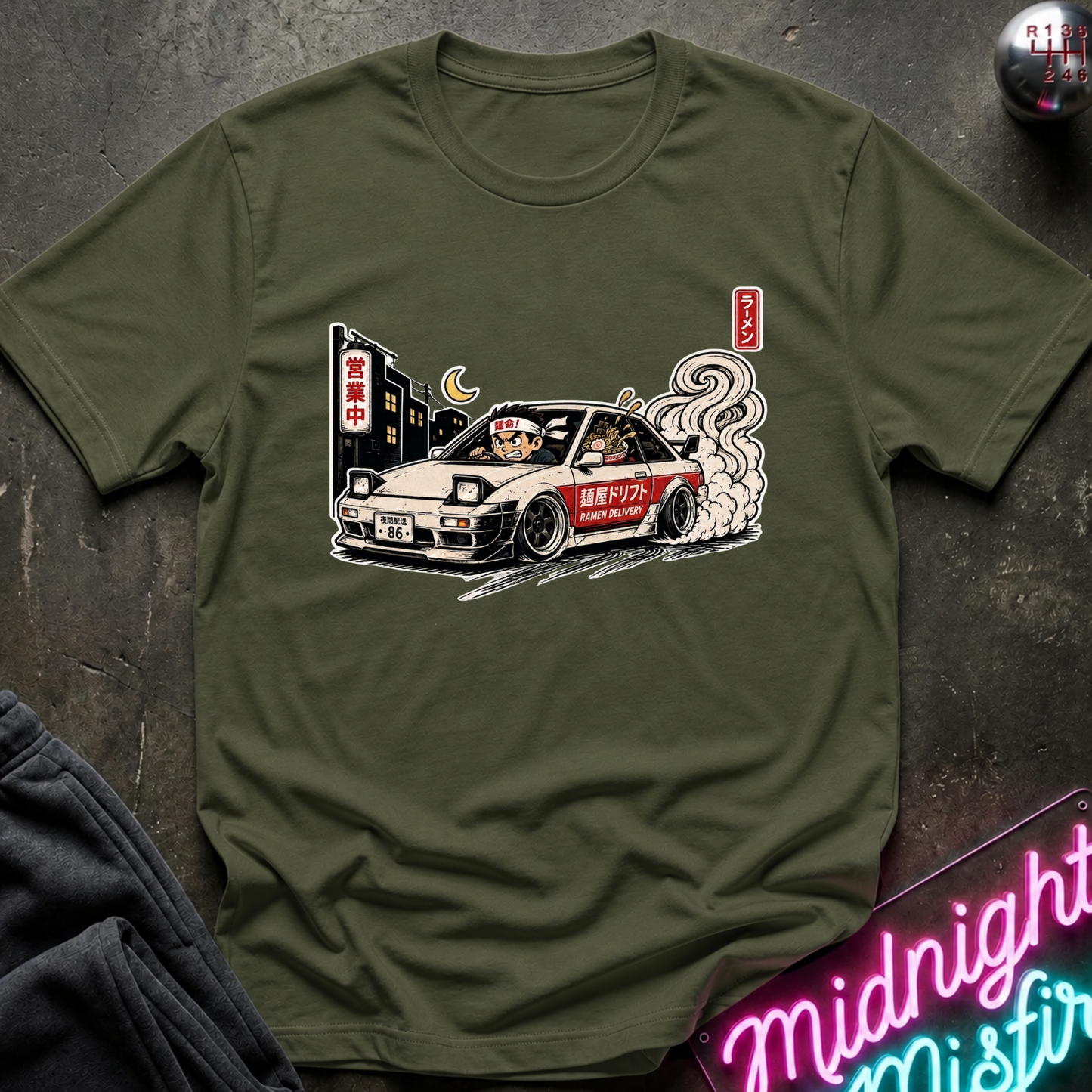 An olive green short-sleeve t-shirt with a graphic print of a white and red race car with smoke, displayed flat on a dark textured surface with a neon sign and dark fabric in the corners.