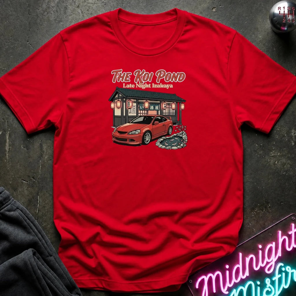 A red short-sleeve graphic t-shirt laid flat on a dark textured surface. The graphic features the text 'THE KOI POND Late Night Hamburger' above an illustration of a red sports car parked in front of a restaurant with red lanterns and a koi pond.