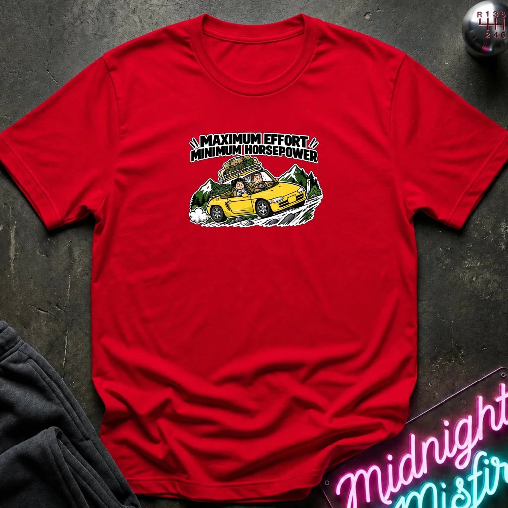 red graphic t-shirt laid flat on a dark textured surface. The graphic features a yellow sports car with a driver and passengers, with the text 'MAXIMUM EFFORT MINIMUM HORSEPOWER' above it.