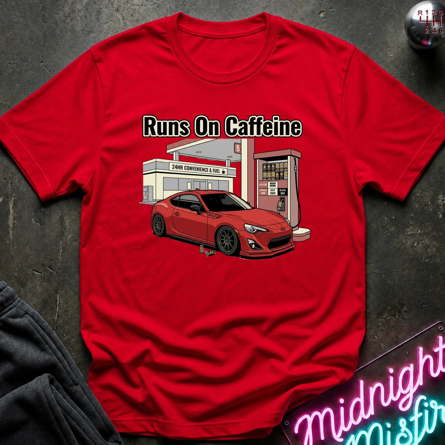 red short-sleeve graphic T-shirt laid flat on a dark textured surface. The shirt features a printed illustration of a red sports car parked in front of a convenience store and gas station, with the text 'Runs On Caffeine' above the graphic.