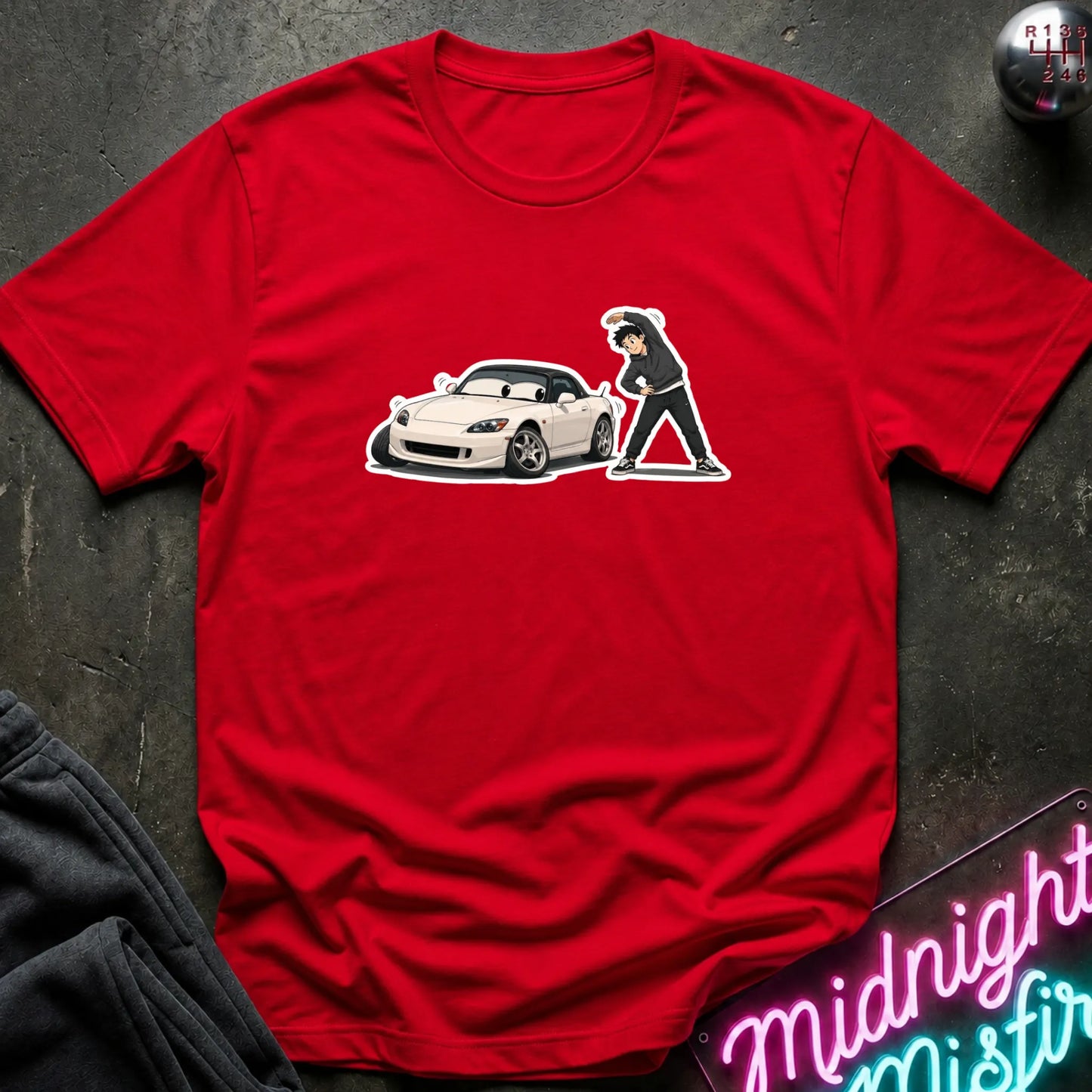 A red short-sleeve crewneck t-shirt laid flat on a dark textured surface. The shirt features a graphic print of a white sports car and a cartoon figure in black and white.