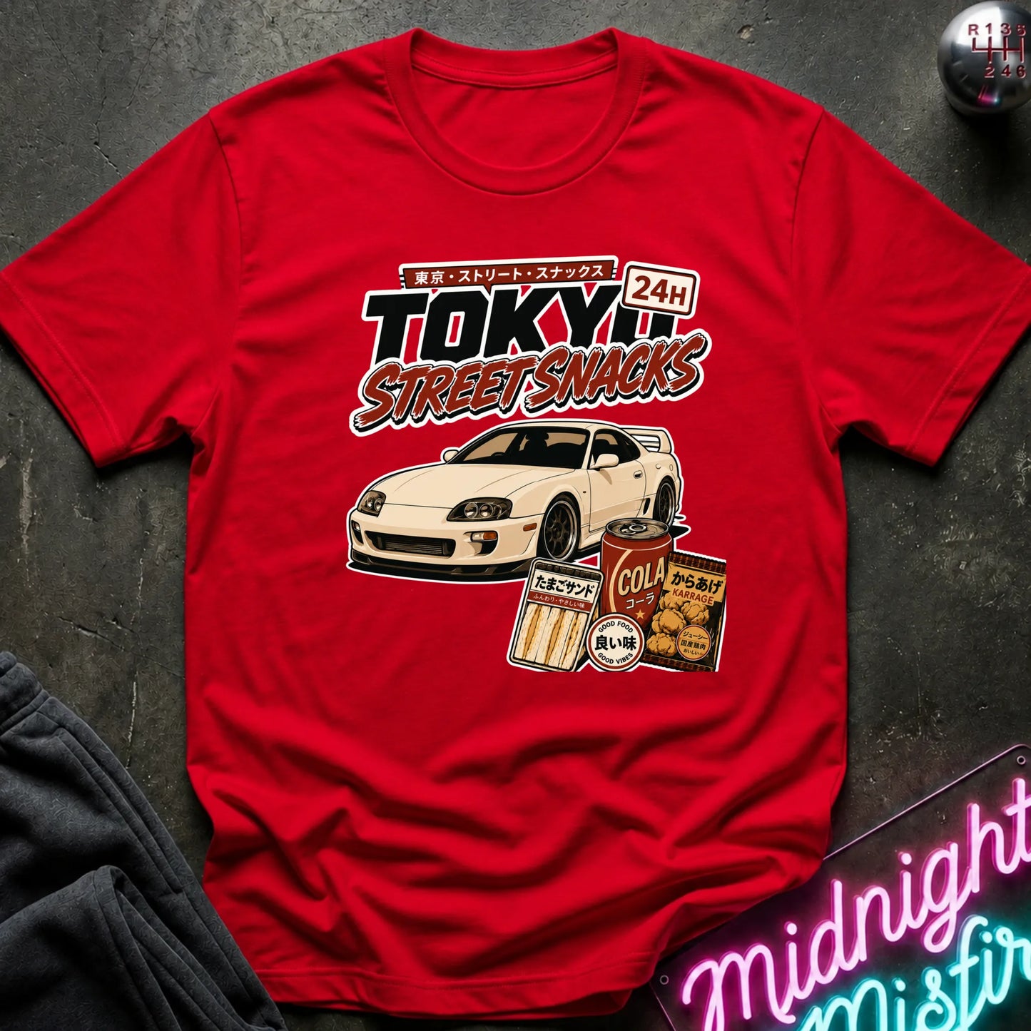 A red short-sleeve graphic T-shirt laid flat on a dark textured surface. The shirt features a large front print with the words “TOKYO STREET SNACKS,” a white sports car, and snack illustrations.
