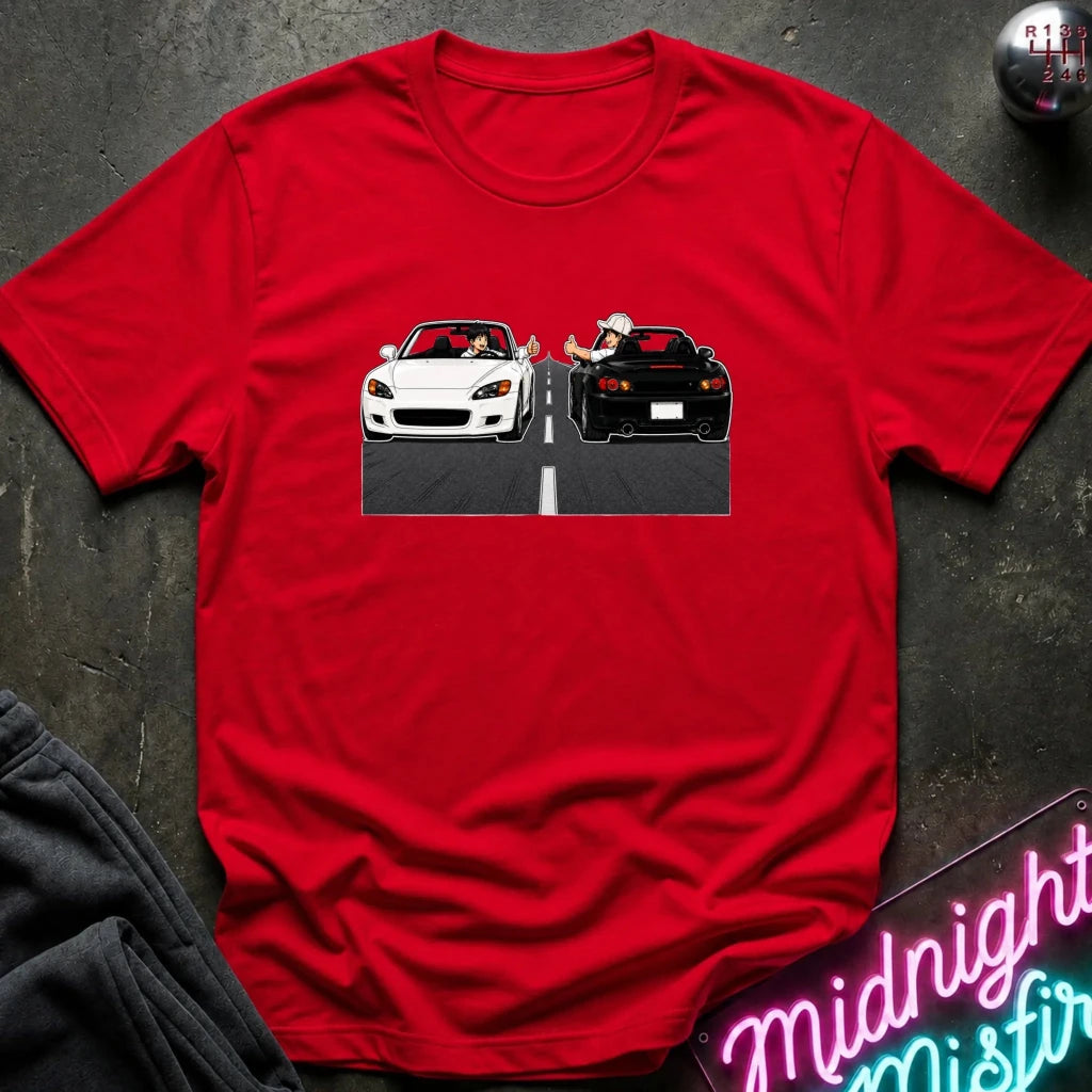 A red short-sleeve crewneck t-shirt laid flat on a dark textured surface. The center of the shirt features a graphic print of a white convertible sports car and a black sports car facing each other on a road with a dashed white line.