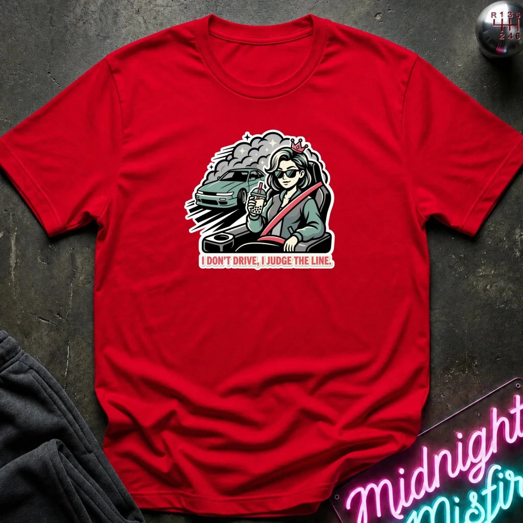 A red short-sleeve crewneck t-shirt laid flat on a dark textured surface. The center of the shirt features a graphic illustration of a woman in a green car with a red seatbelt, with the text 'I DON'T DRIVE, I JUDGE THE LINE.