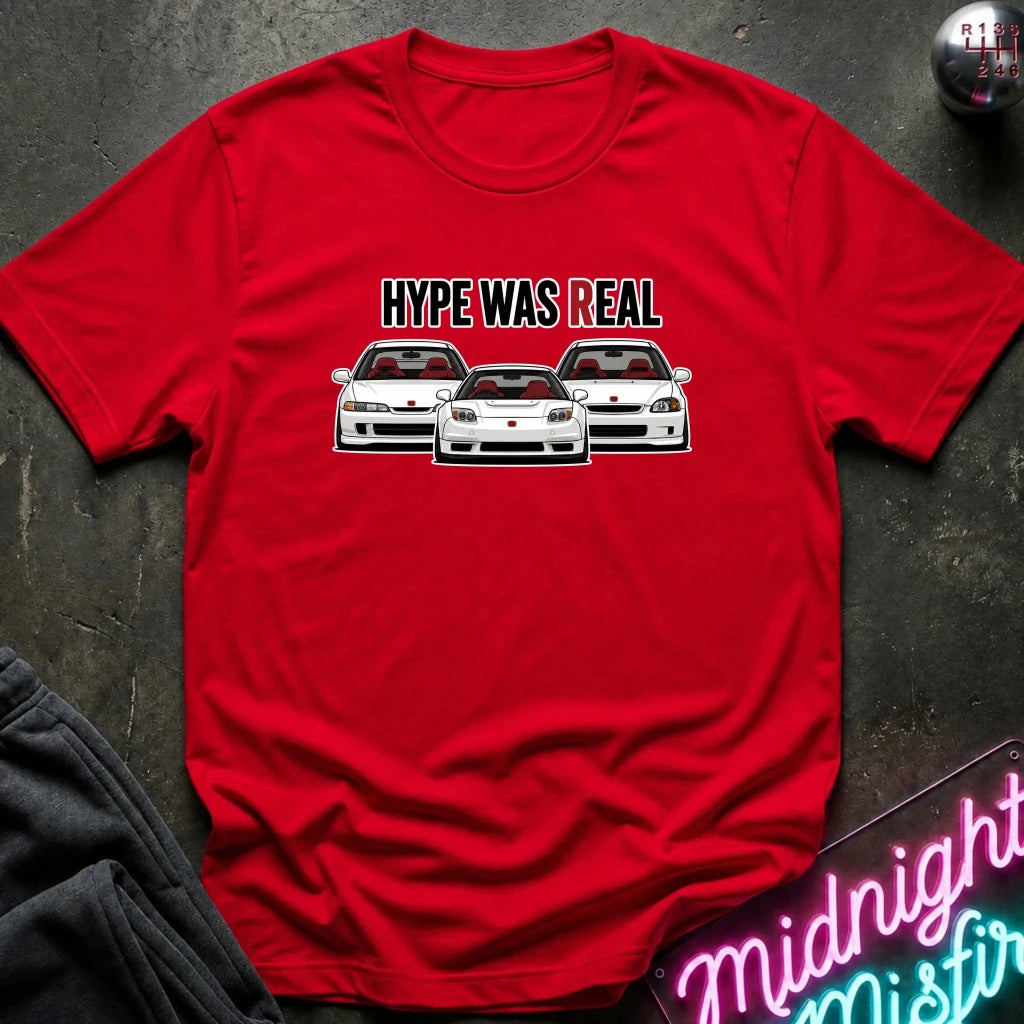 A red short-sleeve crewneck t-shirt laid flat on a dark textured surface. The shirt features a graphic of three white sports cars with the text 'HYPE WAS REAL' above them.