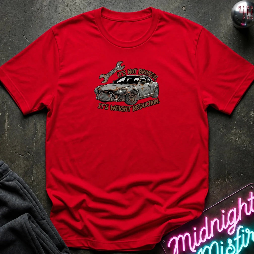 A red short-sleeve crewneck t-shirt laid flat on a dark textured surface. The shirt features a graphic of a silver car with a broken headlight and a wrench, with the text 'IT'S NOT BROKEN' and 'IT'S WEIGHT REDUCTION'.