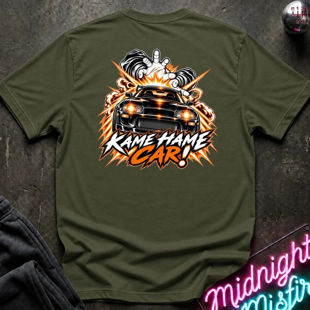 An olive green short-sleeve t-shirt laid flat on a dark textured surface, featuring a large graphic print of a black sports car with flames and the text 'KAME HAME CAR!' in white and orange.