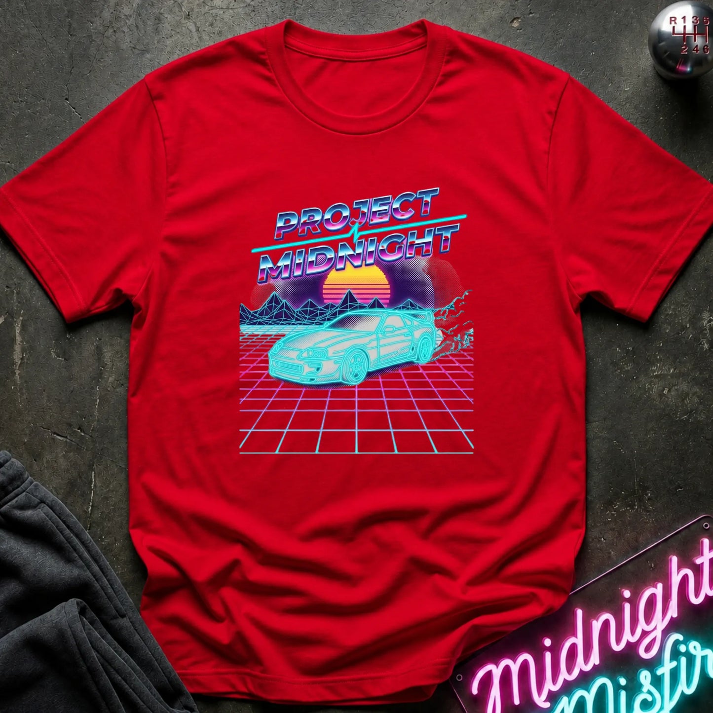 A red short-sleeve t-shirt with a retro 80s synthwave graphic reading 'PROTECT MIDNIGHT' above a neon car and grid scene, laid flat on a dark textured surface with partial jeans and a neon sign visible around it.
