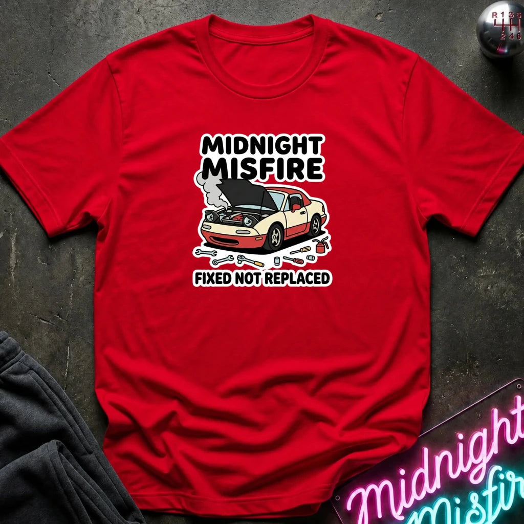 A red short-sleeve graphic T-shirt laid flat on a dark textured surface. The shirt features a printed design of a beige car with its hood open, surrounded by tools, with the text 'MIDNIGHT MISFIRE' above and 'FIXED NOT REPLACED' below.