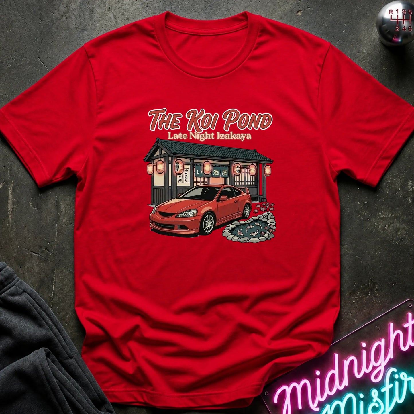 A red short-sleeve graphic t-shirt laid flat on a dark textured surface. The graphic features the text 'THE KOI POND Late Night Hamburger' above an illustration of a red sports car parked in front of a restaurant with red lanterns and a koi pond.