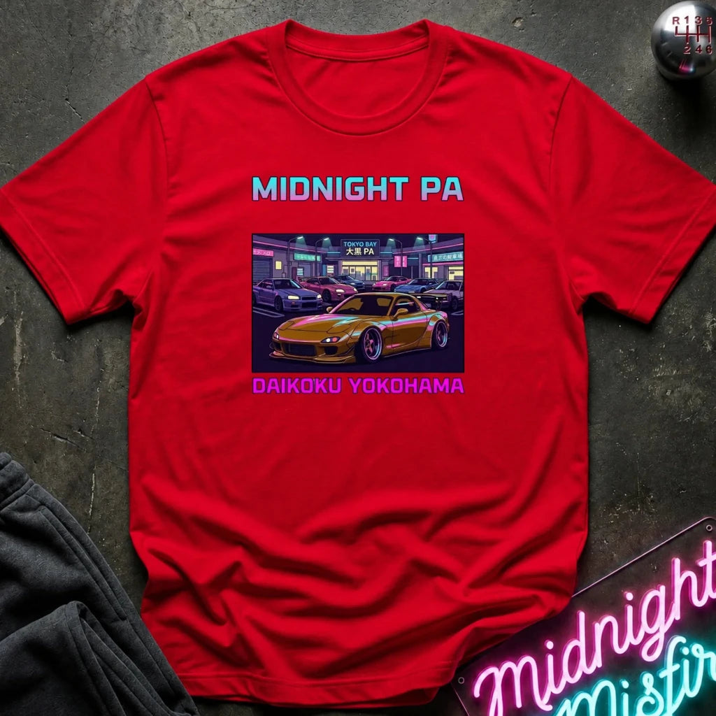 A red short-sleeve graphic T-shirt laid flat on a dark textured surface. The shirt features the text 'MIDNIGHT PA' in light blue at the top and 'DAIKOKU YOKOHAMA' in light pink at the bottom, surrounding a rectangular graphic of a yellow sports car parked in a neon-lit street scene.