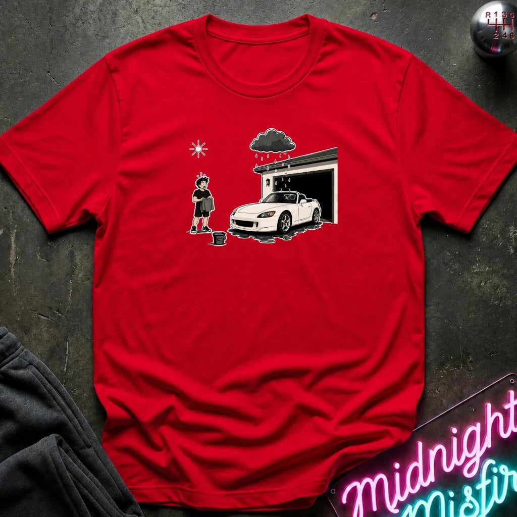 A red short-sleeve crewneck t-shirt laid flat on a dark textured surface. The center of the shirt features a graphic print of a white sports car parked in front of a garage with a person standing nearby.