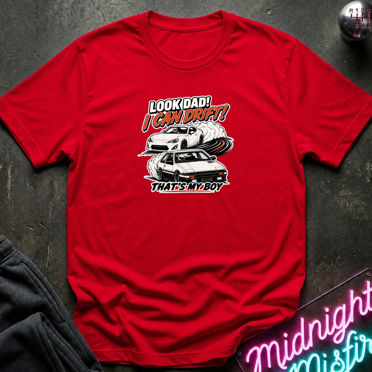A red  short-sleeved t-shirt  that has a cars drifting which are older and newer generations of toyotas.