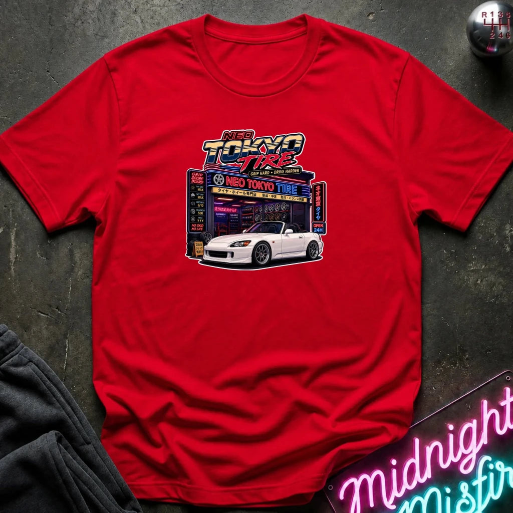 A red short-sleeve crewneck t-shirt featuring a 'Neo Tokyo Tire' graphic with a white sports car, laid flat on a dark textured surface with a neon sign and dark sweatpants partially visible.