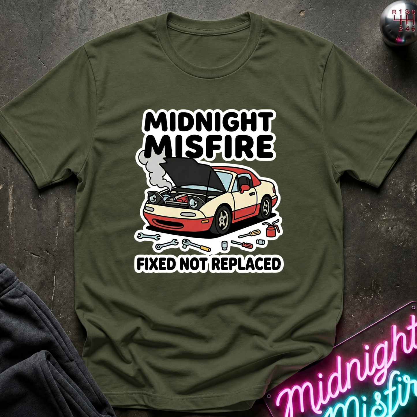 An olive green graphic t-shirt laid flat on a dark textured surface. The shirt features a cartoon illustration of a beige and red sports car with its hood open, surrounded by tools, with the text 'MIDNIGHT MISFIRE' above and 'FIXED NOT REPLACED' below.