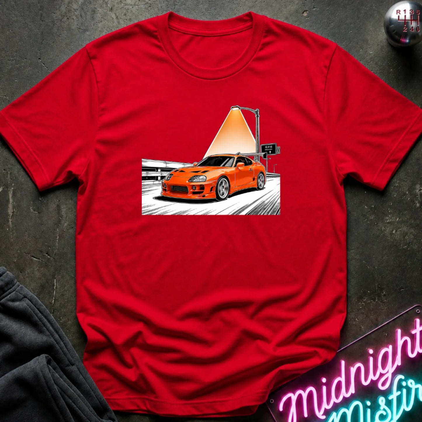 A red short-sleeve t-shirt laid flat on a dark textured surface, featuring a graphic print of an orange sports car speeding on highway.
