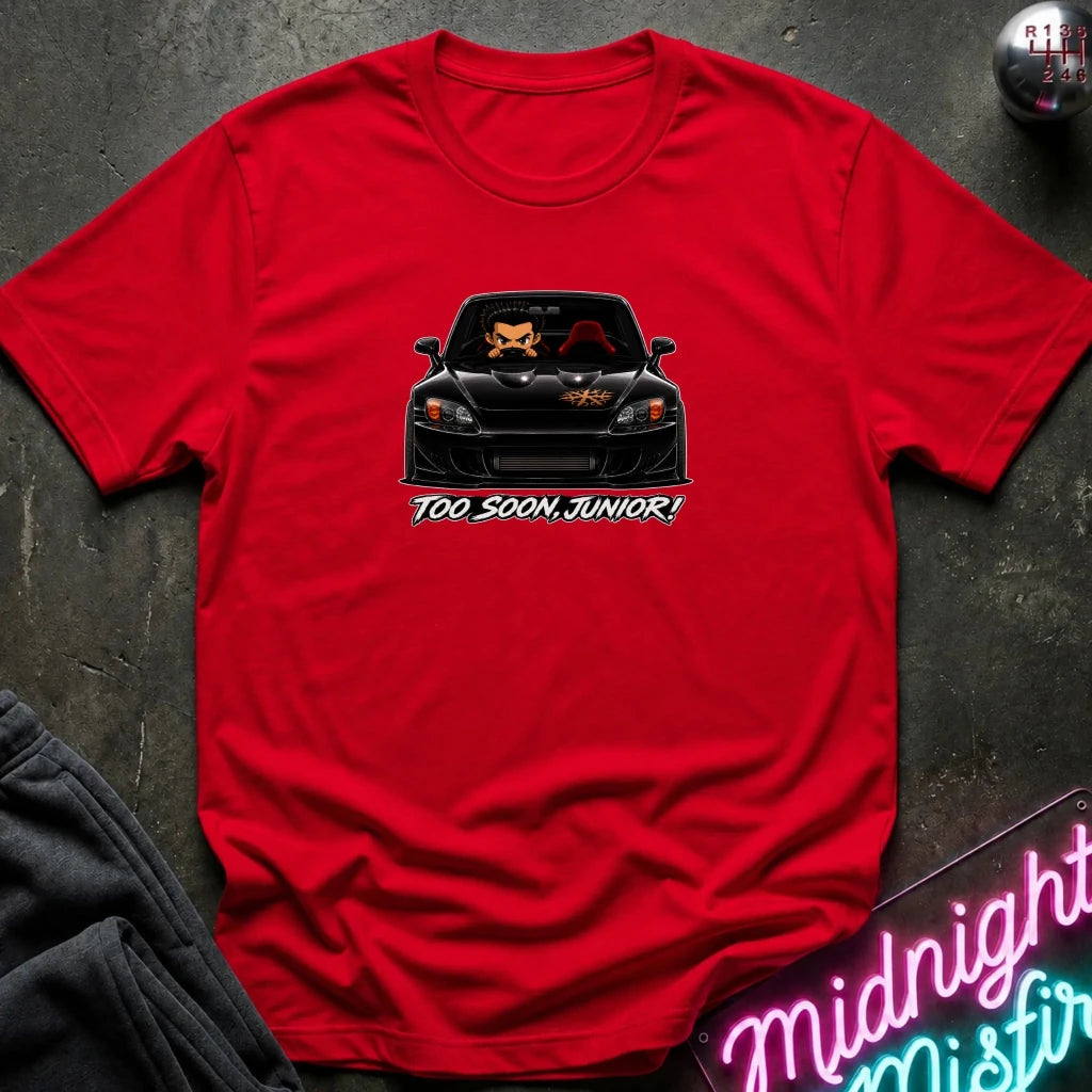 A red short-sleeve crewneck t-shirt laid flat on a dark textured surface. The center of the shirt features a graphic of a black sports car with two cartoon foxes inside, above the text 'TOO SOON, JUNIOR!'.
