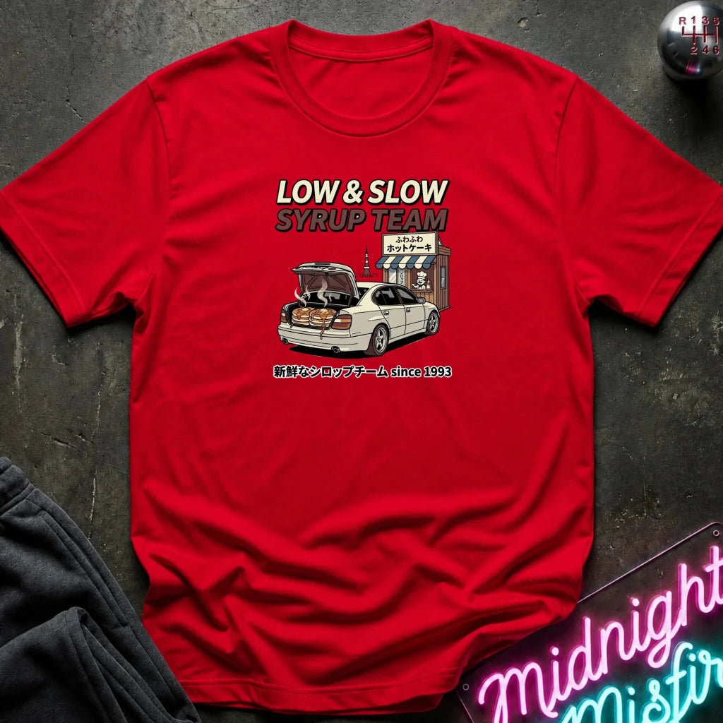 A red short-sleeve graphic t-shirt laid flat on a dark textured surface. The graphic features the text 'LOW & SLOW SYRUP TEAM' above an illustration of a white sedan with its trunk open, parked in front of a Japanese-style shop.