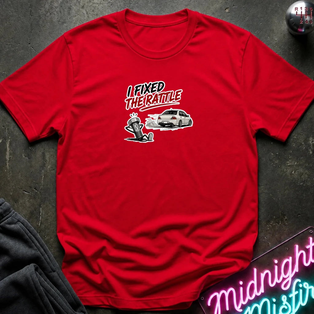 A red short-sleeve graphic t-shirt laid flat on a dark textured surface. The graphic features the text 'I FIXED THE RATLE' in white and red, above a white sports car 