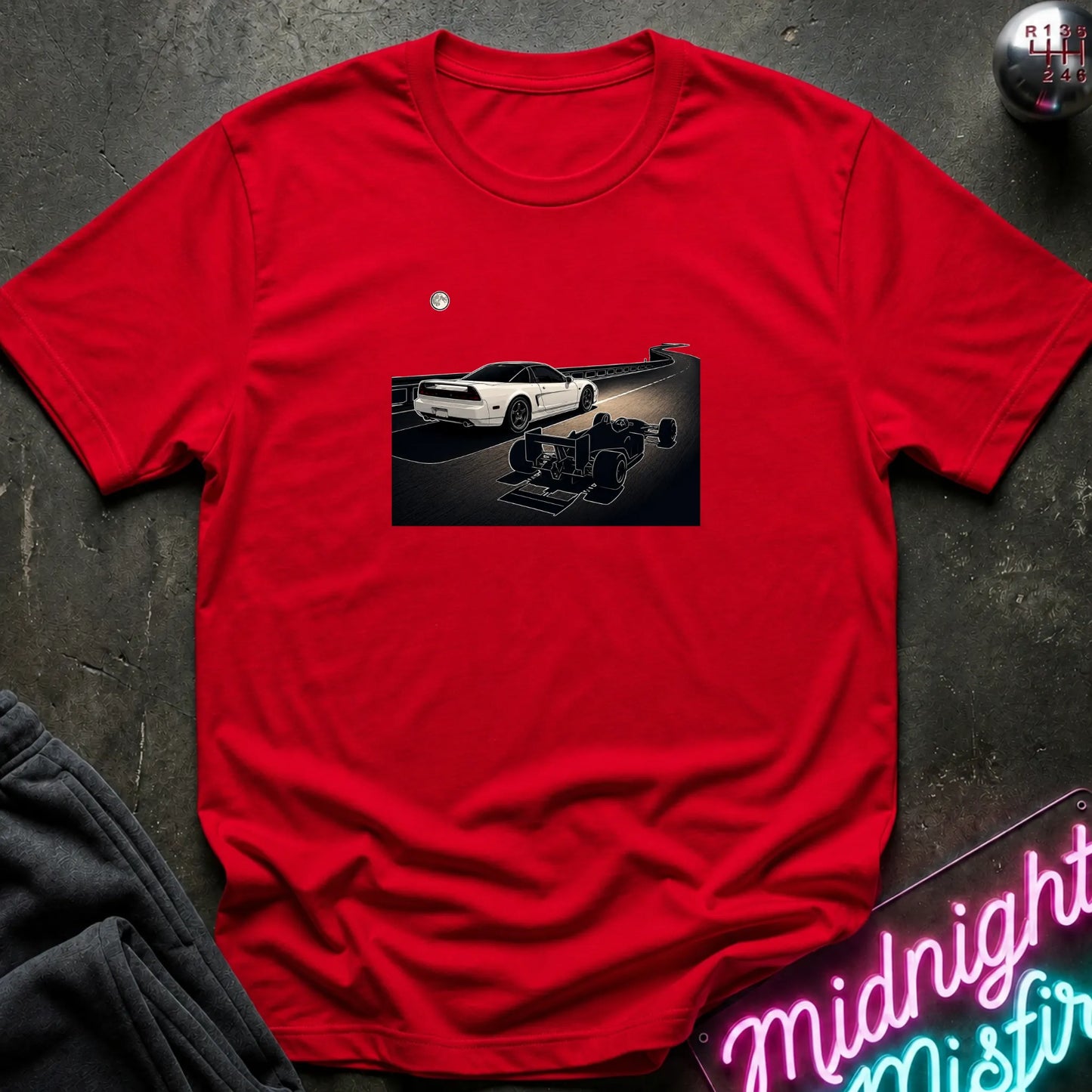 A bright red short-sleeve crewneck t-shirt laid flat on a dark textured surface, featuring a graphic print of a white sports car and a shadow car on a night road scene. The shirt is styled with dark gray sweatpants, a silver ball, and a neon sign in the corners.