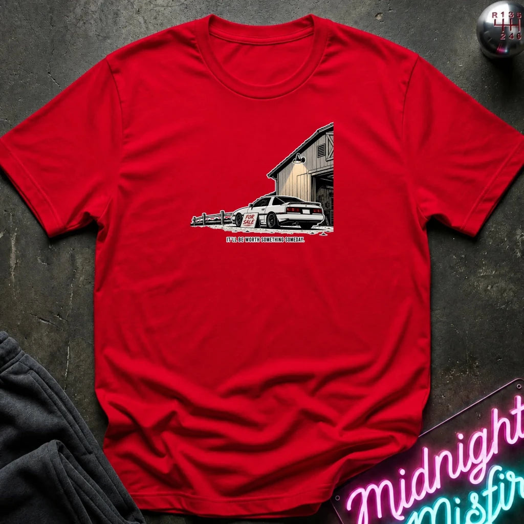 A red short-sleeve crewneck t-shirt laid flat on a dark textured surface, featuring a graphic print of a white sports car parked in front of a garage 